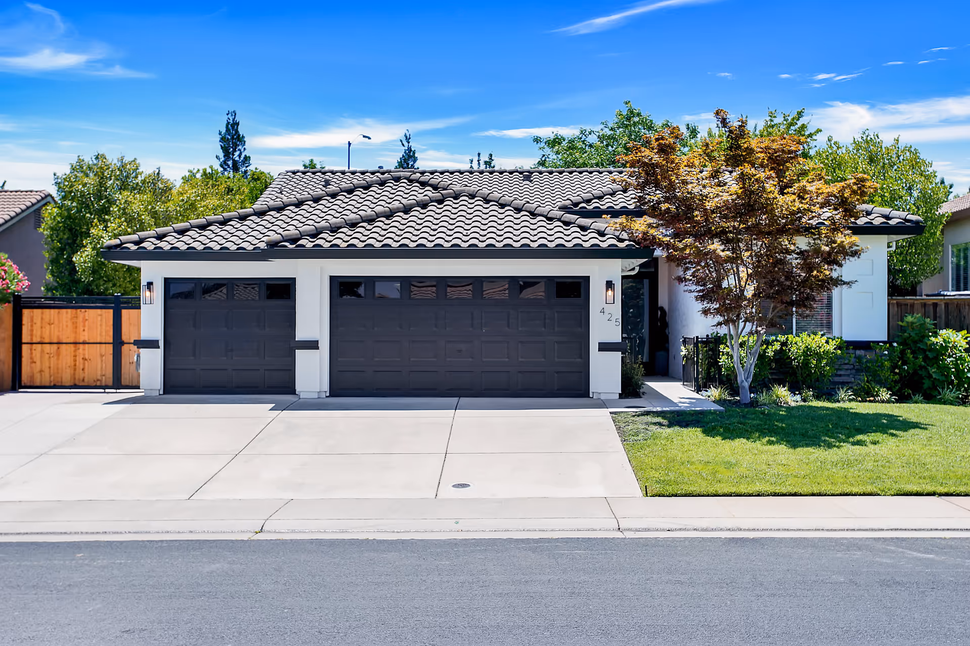 Front exterior view of a single-story house with a tiled roof, two dark garage doors, a driveway, a small tree, and green lawn under a blue sky with some clouds.