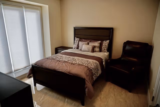 Small bedroom with a made double bed, patterned bedding, a nightstand, leather armchair, and a window with blinds.