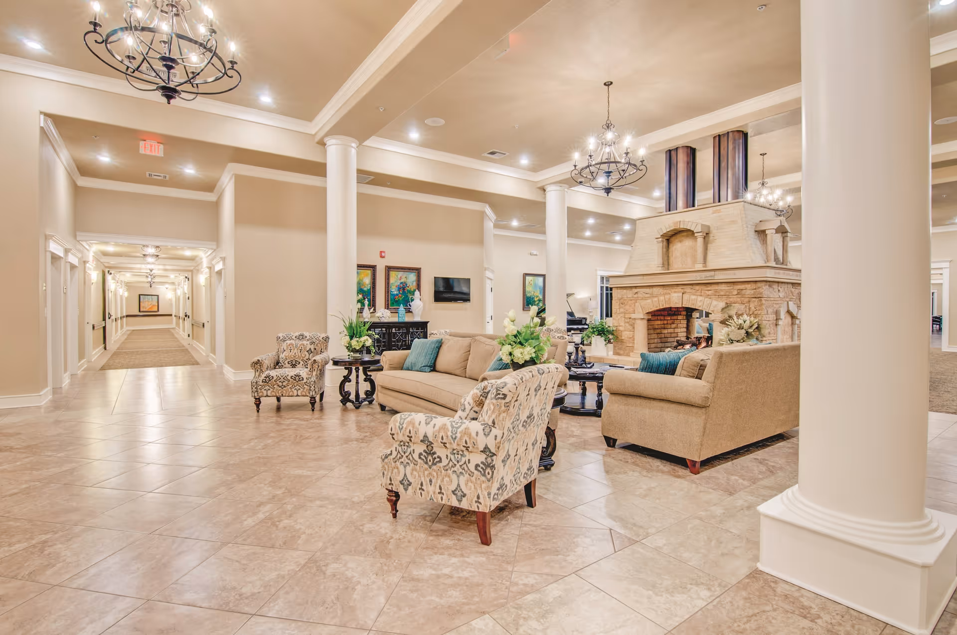 A spacious and elegant living room area in a senior living facility with beige tiled floors, cream-colored walls, and white columns. The room features comfortable beige sofas and patterned armchairs arranged around a large stone fireplace. Decorative chandeliers hang from the ceiling, and there are framed paintings and a flat-screen TV on the walls. A long hallway with additional lighting and artwork extends from the living area.