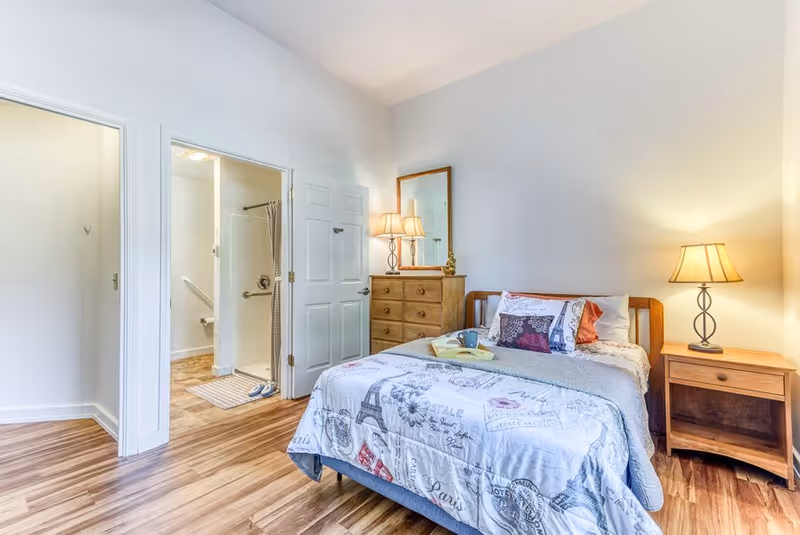 A cozy bedroom in a senior living facility featuring a bed with patterned bedding, a wooden nightstand with a lamp, a wooden dresser with a mirror and lamp, and an open door leading to a bathroom with a shower and grab bars.