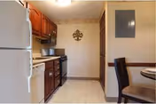 Interior view of a kitchen with wooden cabinets, a white refrigerator, dishwasher, stove, and microwave. The floor has a light-colored tile pattern, and there is a dining table with chairs partially visible on the right side. A decorative wall piece is mounted on the far wall.
