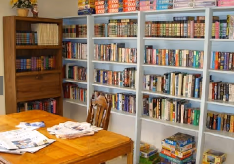 A cozy room with wooden bookshelves filled with books and board games. A wooden table with newspapers and magazines is in the foreground, accompanied by a wooden chair. The room has a warm and inviting atmosphere.