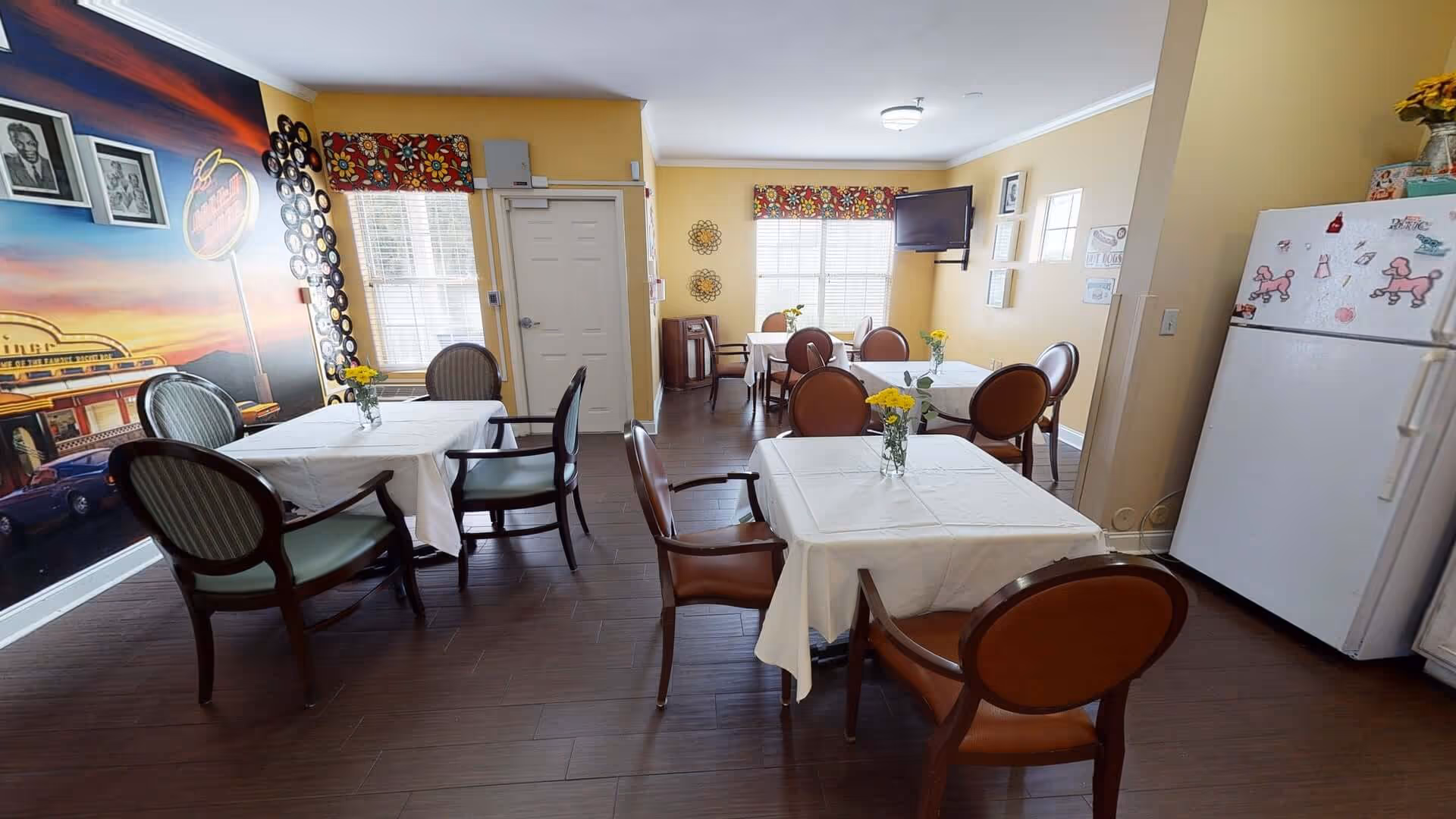 A cozy dining room with several tables covered in white tablecloths, each adorned with a small vase of yellow flowers. The room has yellow walls, a dark wooden floor, and a large mural on one wall depicting a retro diner scene. There are multiple chairs around the tables, a white refrigerator with pink poodle magnets, and a wall-mounted TV near a window with floral valances.