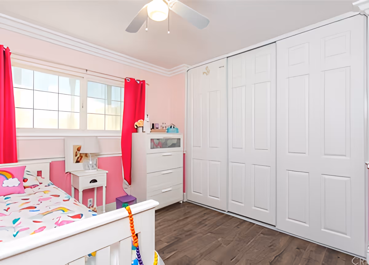 A bright bedroom with pink walls and white trim, featuring a white bed with colorful bedding including rainbows and hearts, a white nightstand with a lamp, a white dresser, large white closet doors, a ceiling fan with a light, and red curtains on the window.