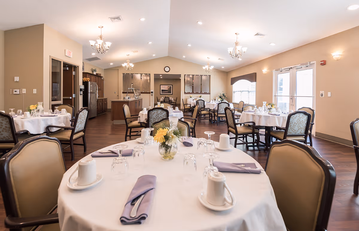 A bright and spacious dining room in a senior living facility with round tables covered in white tablecloths, each set with cups, glasses, silverware wrapped in lavender napkins, and small floral centerpieces. The room features wooden flooring, beige walls, chandeliers, and large windows allowing natural light to enter. There is a kitchen area visible in the background.