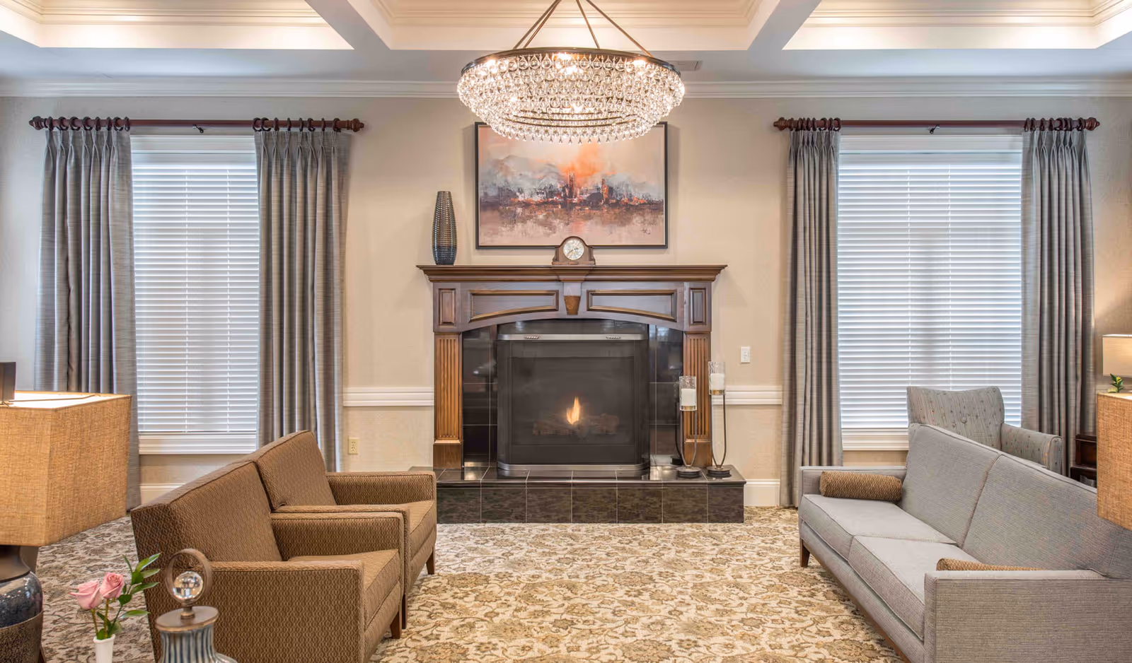 A cozy living room with a lit fireplace framed by a wooden mantel. Above the mantel is a colorful abstract painting. Two large windows with gray curtains flank the fireplace. The room has a patterned carpet and is furnished with a brown loveseat on the left, a gray sofa on the right, and an armchair in the corner. A large, elegant chandelier hangs from the ceiling.