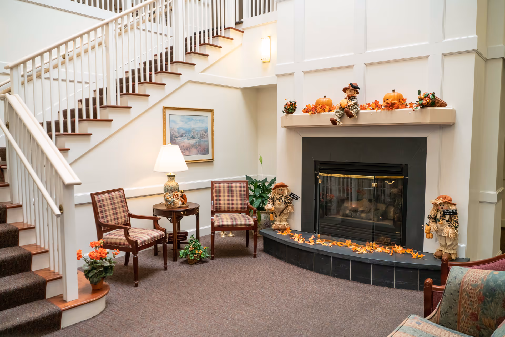 A cozy interior common area featuring a staircase with white railings, two plaid upholstered chairs around a small wooden table with a lamp, and a fireplace decorated with autumn-themed items including pumpkins, scarecrows, and fall leaves.