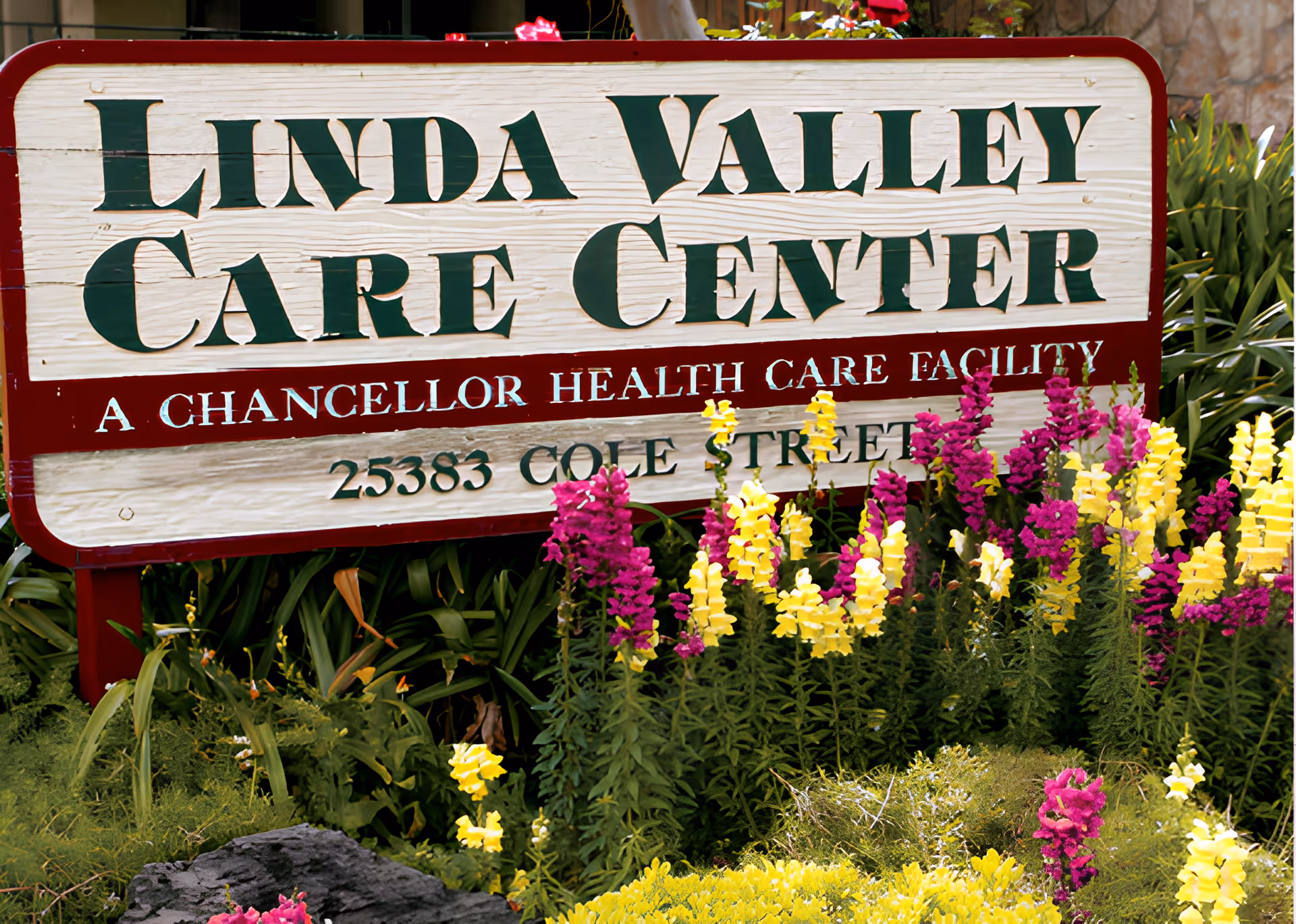 A wooden sign for Linda Valley Care Center, a Chancellor Health Care Facility, surrounded by colorful yellow and purple flowers and greenery.