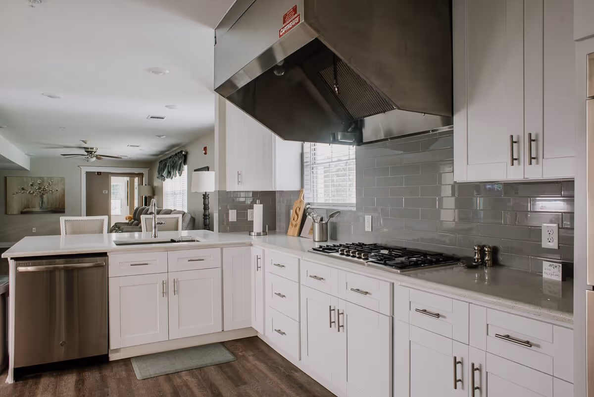 Modern kitchen with white cabinets, a stainless steel dishwasher, a gas stove with a large stainless steel range hood, and a white countertop. The kitchen opens into a living area with a ceiling fan, a lamp, and a painting on the wall.