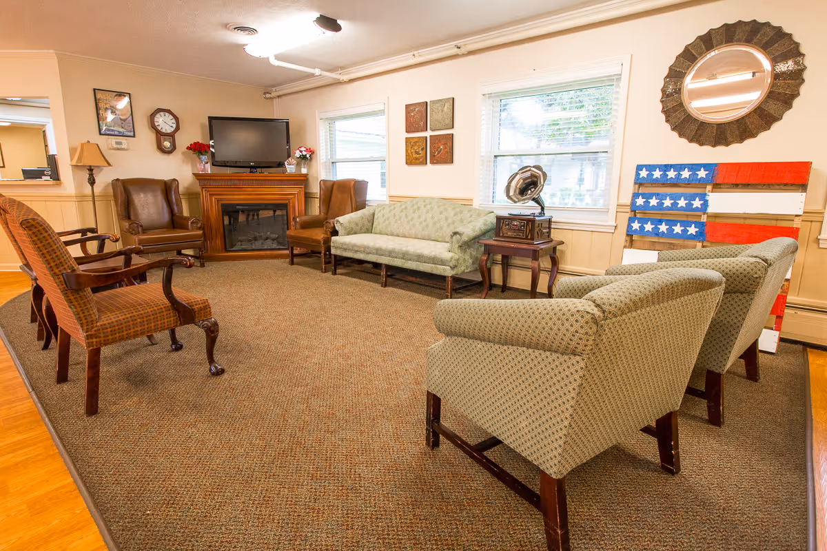 A cozy living room with a variety of upholstered chairs and sofas arranged around a carpeted area. There is a wooden fireplace with a TV mounted above it, two windows letting in natural light, and decorative items including a vintage gramophone on a side table, a round mirror, and a wooden American flag wall decoration.