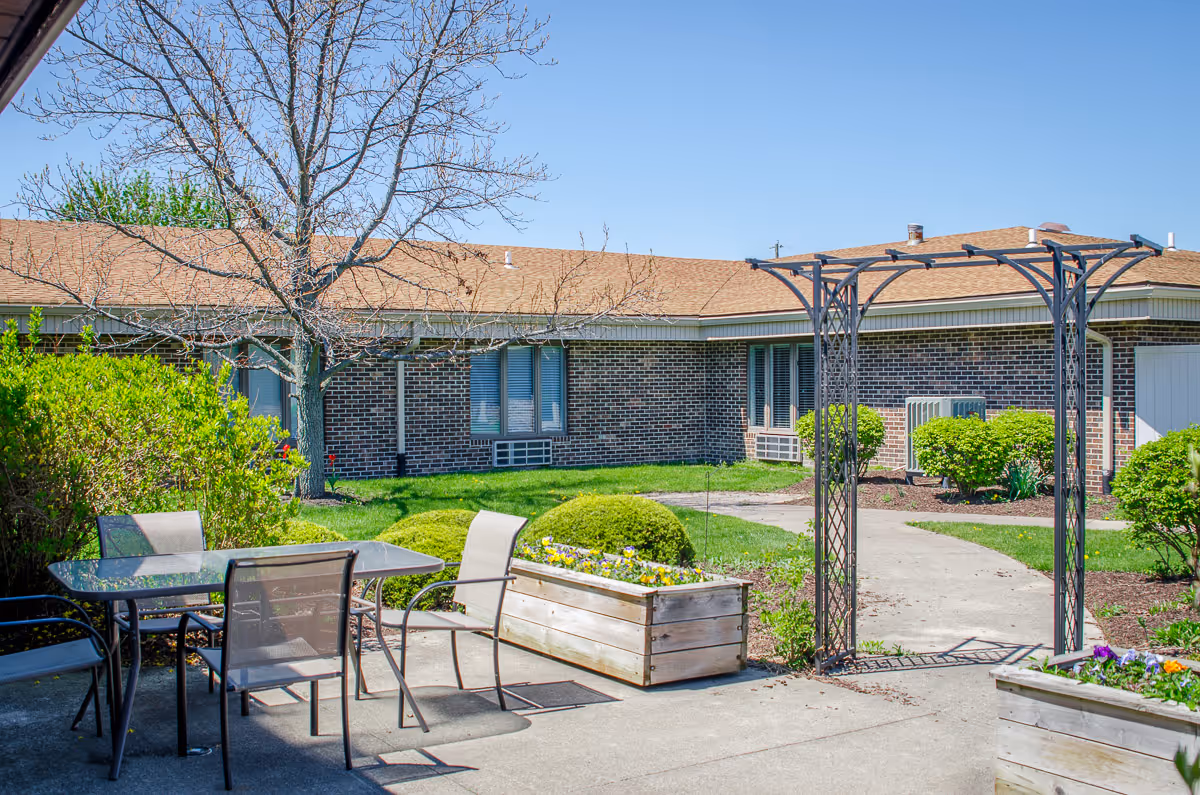 Outdoor patio area with a glass table and four chairs, surrounded by greenery including bushes and a leafless tree. There is a metal archway and a brick building with windows in the background under a clear blue sky.