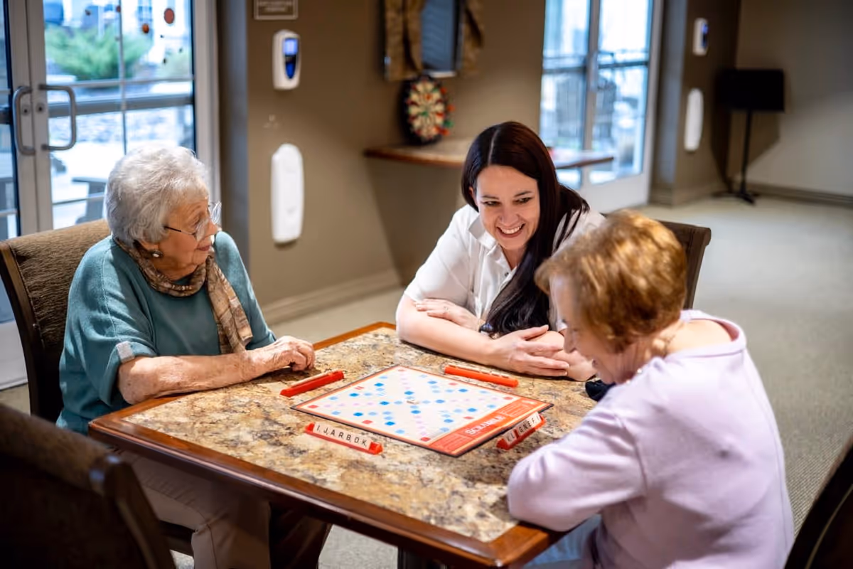 Two elderly women and a younger woman sitting around a table playing a board game in a well-lit room with large windows and a dartboard on the wall.