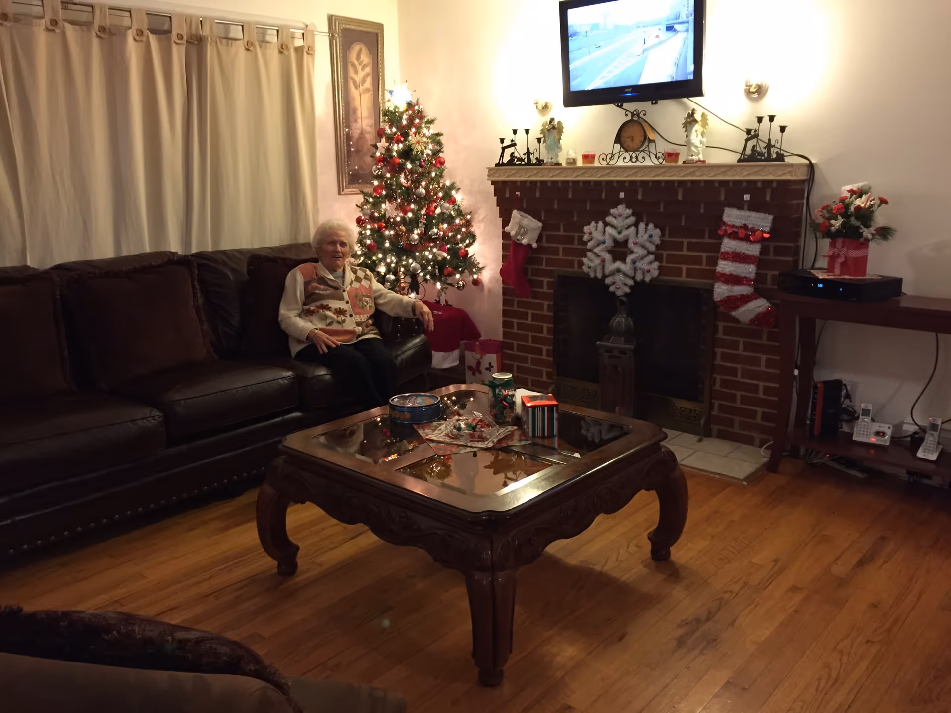 A cozy living room decorated for Christmas with a Christmas tree lit with white and red lights, a brick fireplace adorned with stockings and a large snowflake decoration, and a mounted TV above the fireplace. An elderly woman is sitting on a dark brown leather sofa. A wooden coffee table with holiday-themed items is in the center of the room, and there is a side table with a flower arrangement and electronic devices.