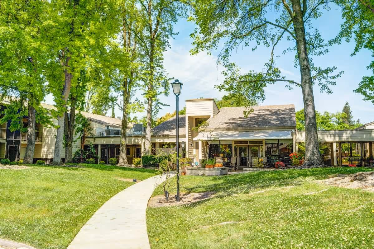 A sunny outdoor view of Greenhaven Place featuring a paved walkway leading through a grassy area with tall trees and a lamppost. The building has large windows and a covered patio area with seating, surrounded by greenery and flowers.