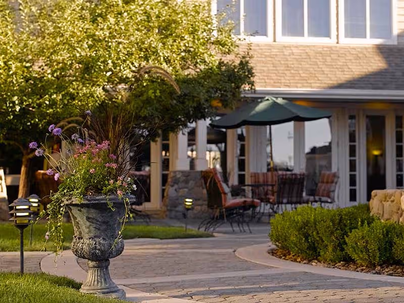 Outdoor patio area with a stone pathway, a large decorative planter with flowers and grasses, green bushes, and outdoor seating with cushions under a green umbrella in front of a building with large windows.
