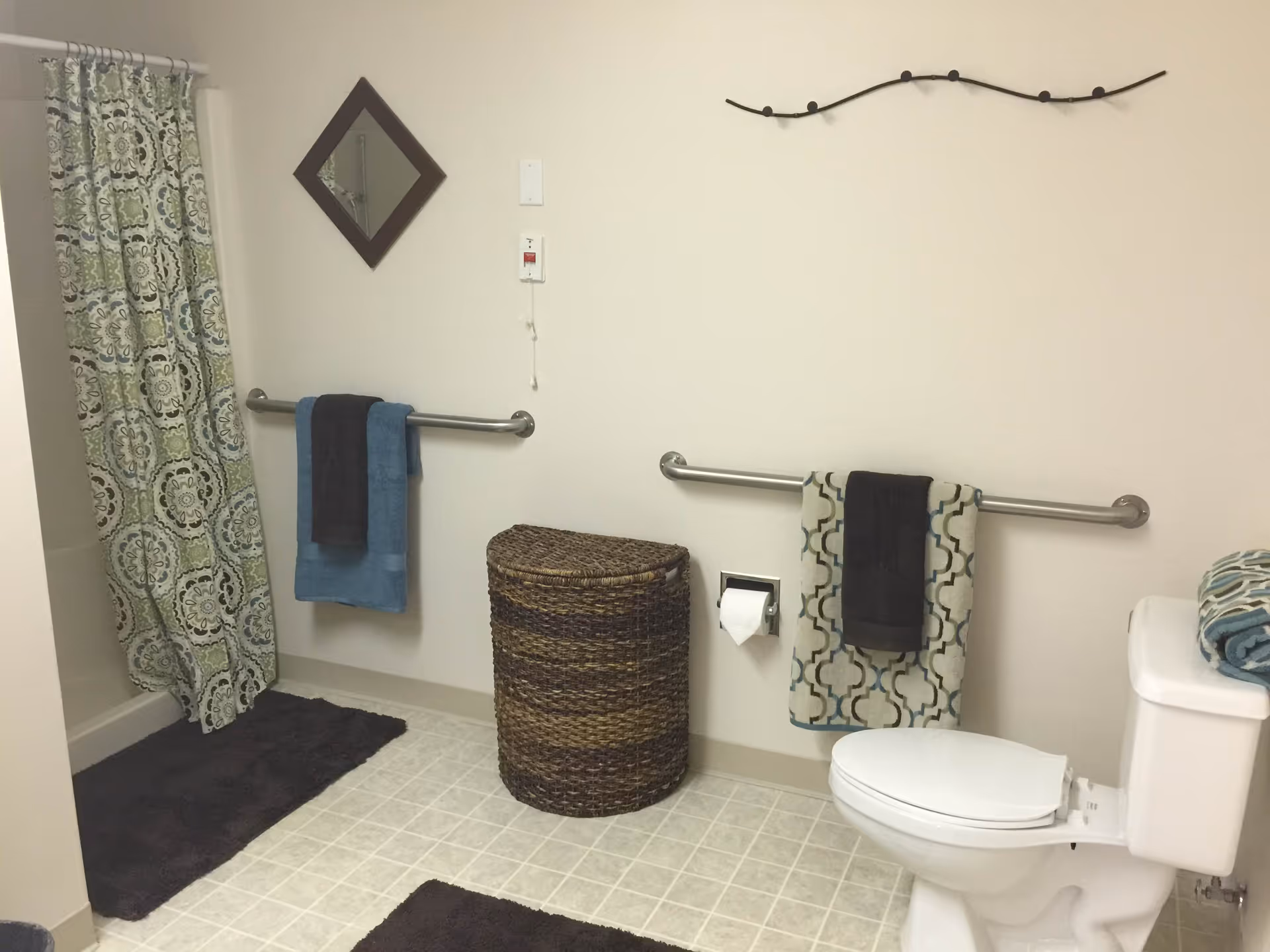 A bathroom with a white toilet, a woven laundry basket, two towel bars with towels, a patterned shower curtain, a small diamond-shaped mirror on the wall, and a decorative wall hook above the toilet.