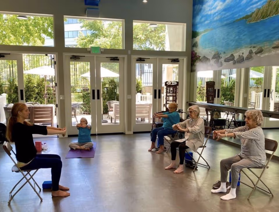 A group of elderly women and a young instructor participate in a seated exercise class in a bright room with large windows and glass doors leading to an outdoor patio. The women are seated on chairs arranged in a semi-circle, stretching their arms forward. The room has a mural of a beach scene on one wall and mirrors on another.