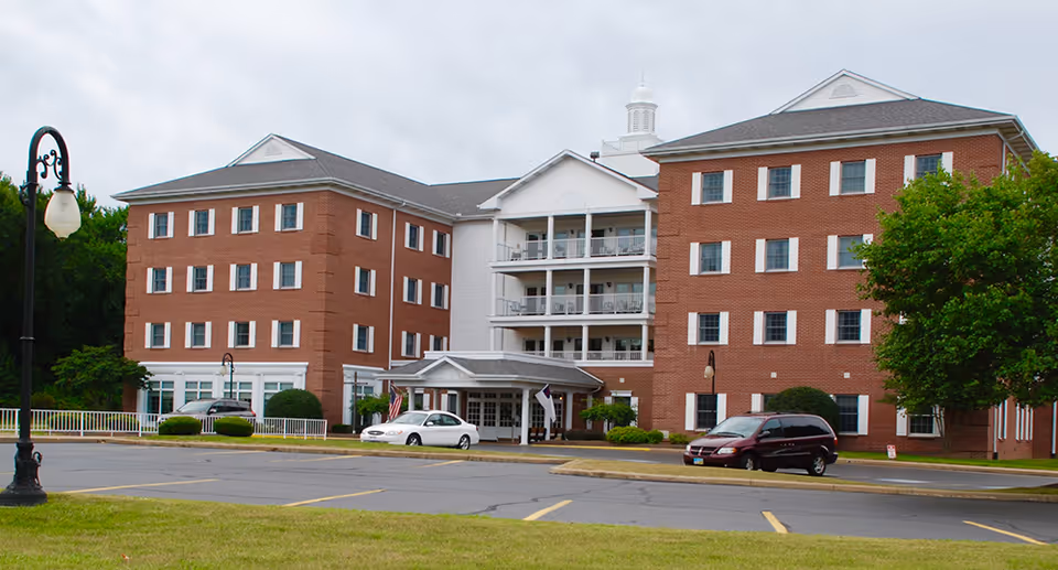 Exterior view of a multi-story brick senior living facility with white trim and a covered entrance. There are several cars parked in front and a street lamp on the left side. The building is surrounded by greenery and trees under a cloudy sky.