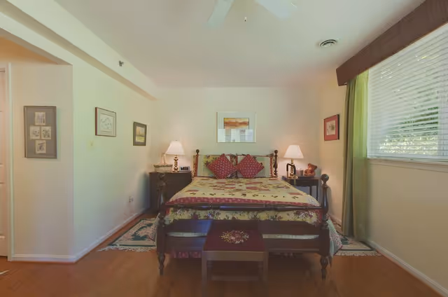 A cozy bedroom with a wooden bed featuring a floral quilt and three decorative pillows. There are two bedside tables with lamps on each side of the bed. The room has light-colored walls adorned with framed pictures, a window with green curtains, and a ceiling fan above. A small wooden bench is placed at the foot of the bed on a hardwood floor.