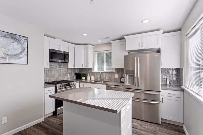 A modern kitchen with white cabinets, stainless steel appliances including a refrigerator, microwave, and stove. There is a kitchen island with a smooth countertop in the center, a window above the sink, and a piece of abstract art on the wall.