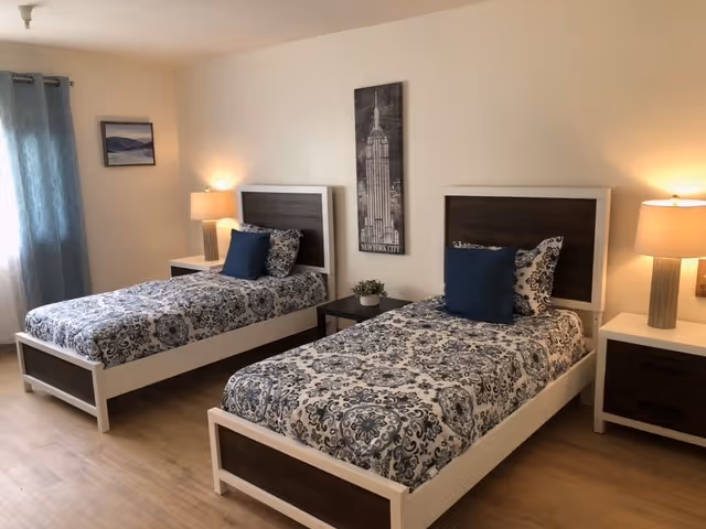 A bedroom with two single beds featuring white frames and dark wood headboards. Each bed is made with patterned blue and white bedding and has a dark blue pillow. Between the beds is a small dark wood nightstand with a small plant on it. On either side of the beds are matching nightstands with table lamps. The room has light-colored walls and wood flooring, with a window covered by blue curtains on the left side. A black and white framed picture of a city building hangs on the wall between the beds.