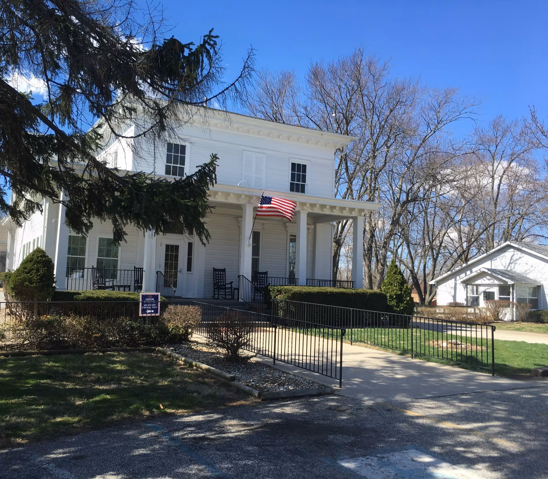 Exterior view of a two-story white building with a front porch featuring rocking chairs and an American flag. There is a wheelchair accessible ramp leading to the entrance, surrounded by bushes and trees with no leaves. A smaller white building is visible in the background under a clear blue sky.