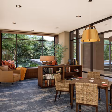 A bright and cozy common area with large windows showing green trees outside. The room features a wooden table with four woven chairs, a bookshelf filled with books, a comfortable armchair with colorful pillows and a blanket, and a modern pendant light hanging from the ceiling.