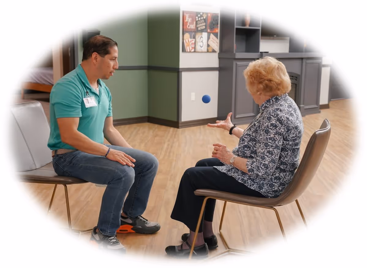 An elderly woman sitting on a chair and tossing a small blue ball to a man sitting opposite her in a room with wooden flooring and green and gray walls.