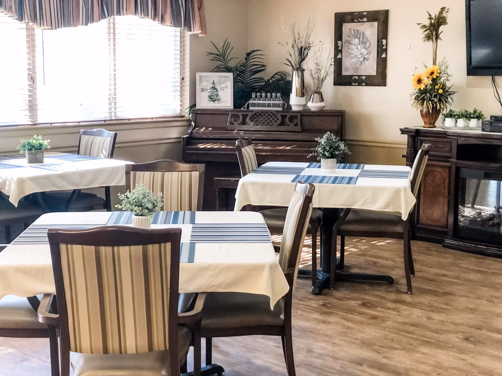 Bright dining room with tables covered in white tablecloths and striped runners, chairs, potted plants, a piano and decorative accents.