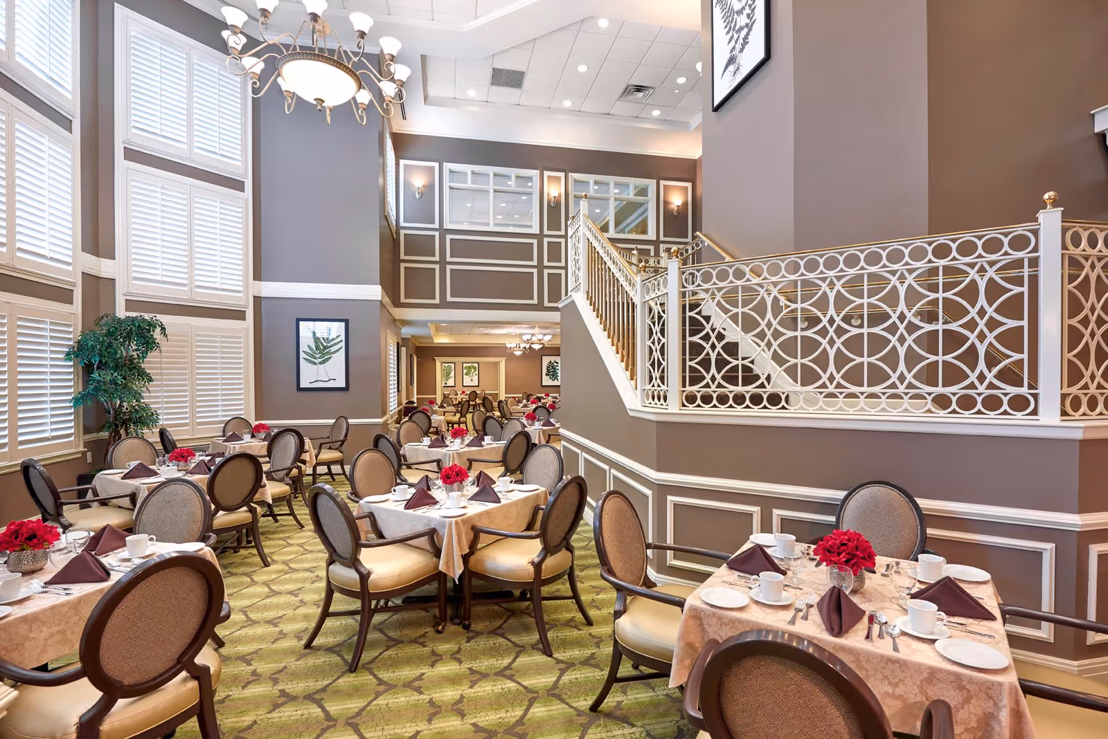 A spacious and elegant dining room with multiple tables set with beige tablecloths, white plates, cups, and silverware. Each table has a centerpiece of red flowers and folded dark napkins. The room features high ceilings, large windows with white shutters, a decorative chandelier, and a staircase with ornate white railing. The walls are painted in a warm taupe color with white trim and framed botanical artwork.