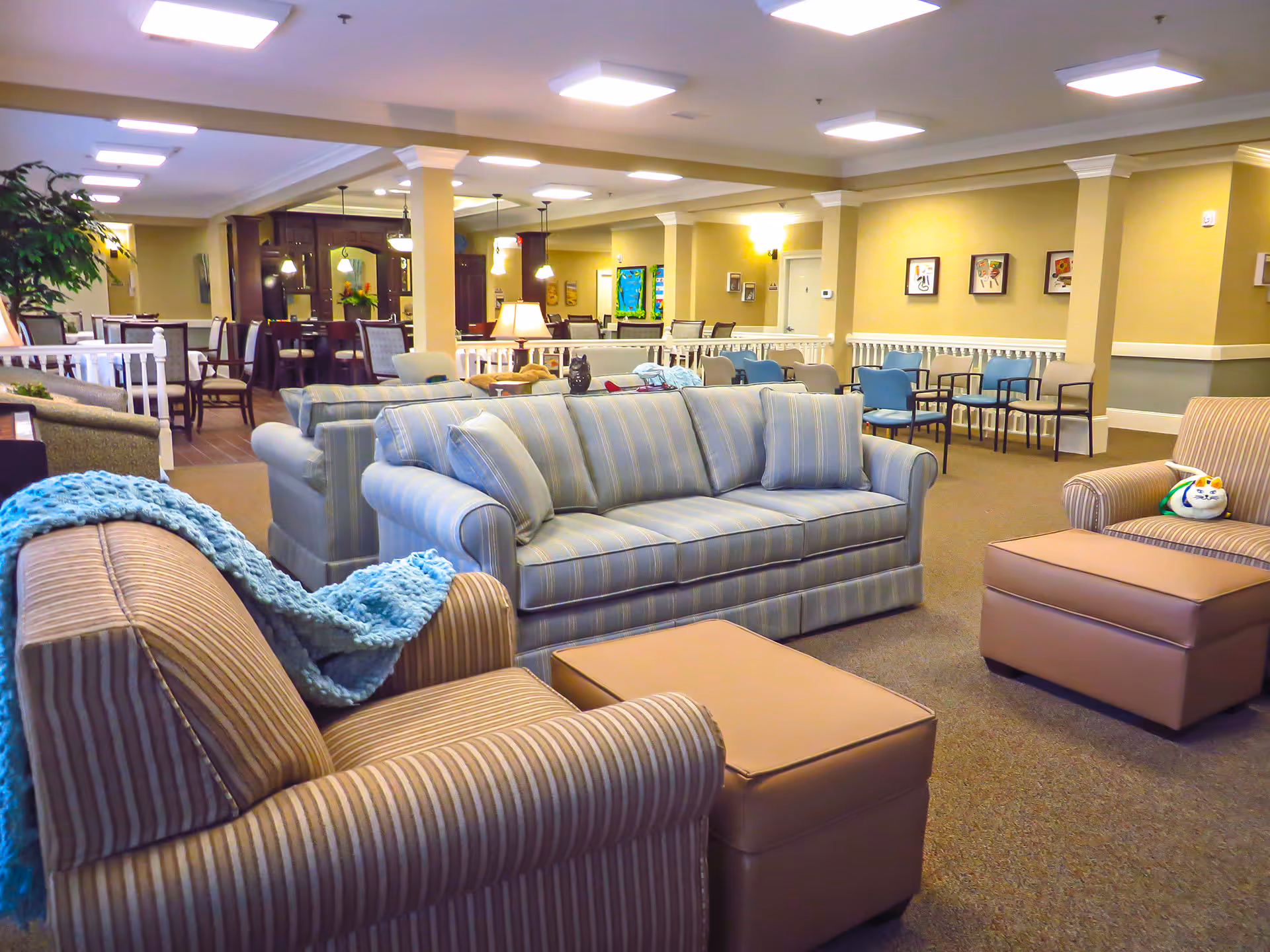 A spacious and well-lit common area in a senior living facility featuring multiple striped sofas and armchairs with cushions, a blue knitted throw blanket, and ottomans. The room has beige walls with framed artwork, carpeted floors, and overhead fluorescent lighting. In the background, there are additional seating areas with chairs and tables, as well as a dining area with dark wood furniture and pendant lights.