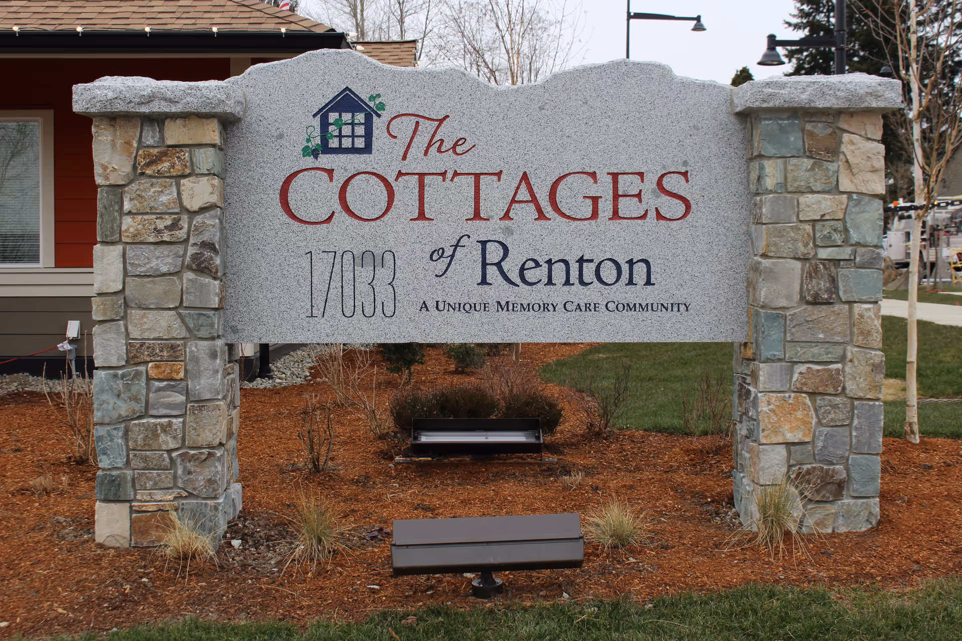 Stone sign for The Cottages of Renton, a unique memory care community, set outdoors with landscaping and a building partially visible in the background.