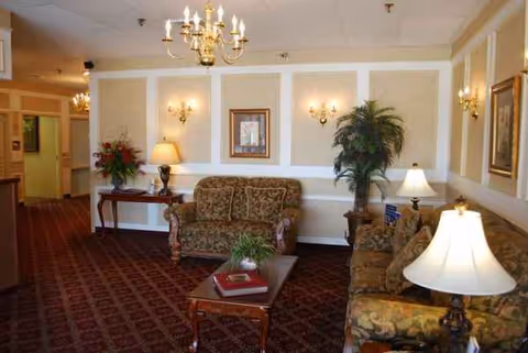 A cozy senior living lounge with patterned sofas, chairs, lamps, and plants.