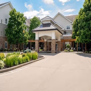 Front exterior view of a senior living facility named Pinebrook with a covered entrance, surrounded by trees and landscaping under a partly cloudy sky.