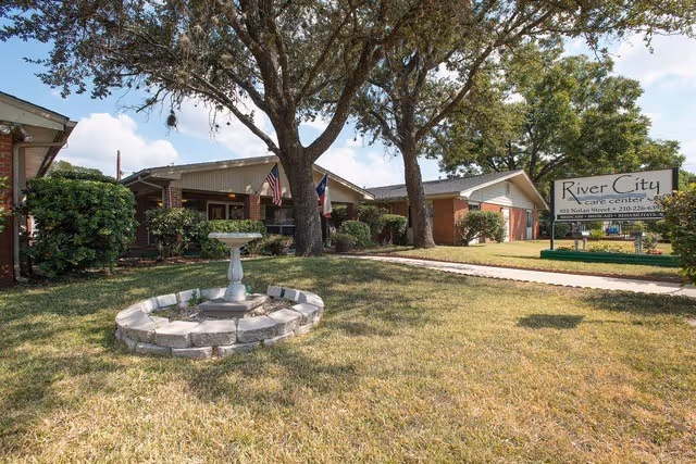 Exterior view of River City Care Center showing a single-story brick building with a covered entrance, surrounded by trees and shrubs. A stone birdbath is in the foreground on a grassy lawn, and a sign with the facility's name and contact information is visible near the sidewalk.
