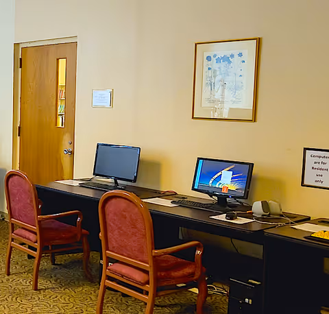 A small computer station with two desktop computers on a long black desk. Two wooden chairs with red cushions are placed in front of the computers. A wooden door with a small window is on the left side of the beige-colored wall. A framed floral artwork hangs on the wall above the desk. A sign on the wall indicates that the computers are for resident use only.