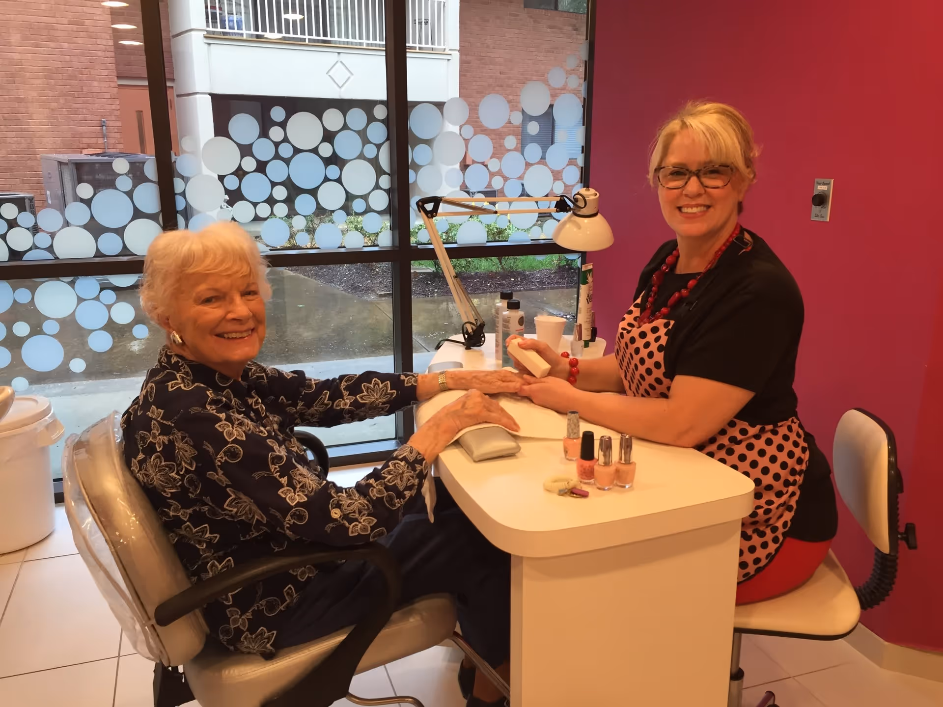 An elderly woman is smiling while getting a manicure from a nail technician in a bright room with large windows decorated with circular frosted patterns. The nail technician is also smiling and wearing glasses and a pink and black polka dot apron. Various nail polish bottles and manicure tools are on the table.