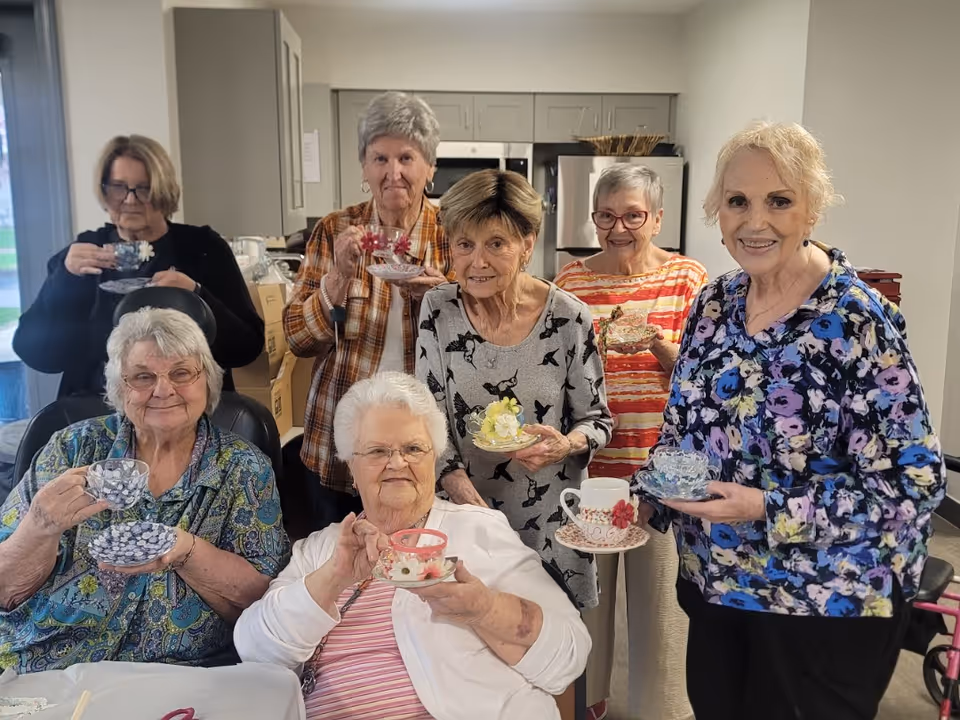 Seven elderly women gathered indoors in a kitchen area, each holding a decorative teacup and saucer, smiling and posing for the photo.
