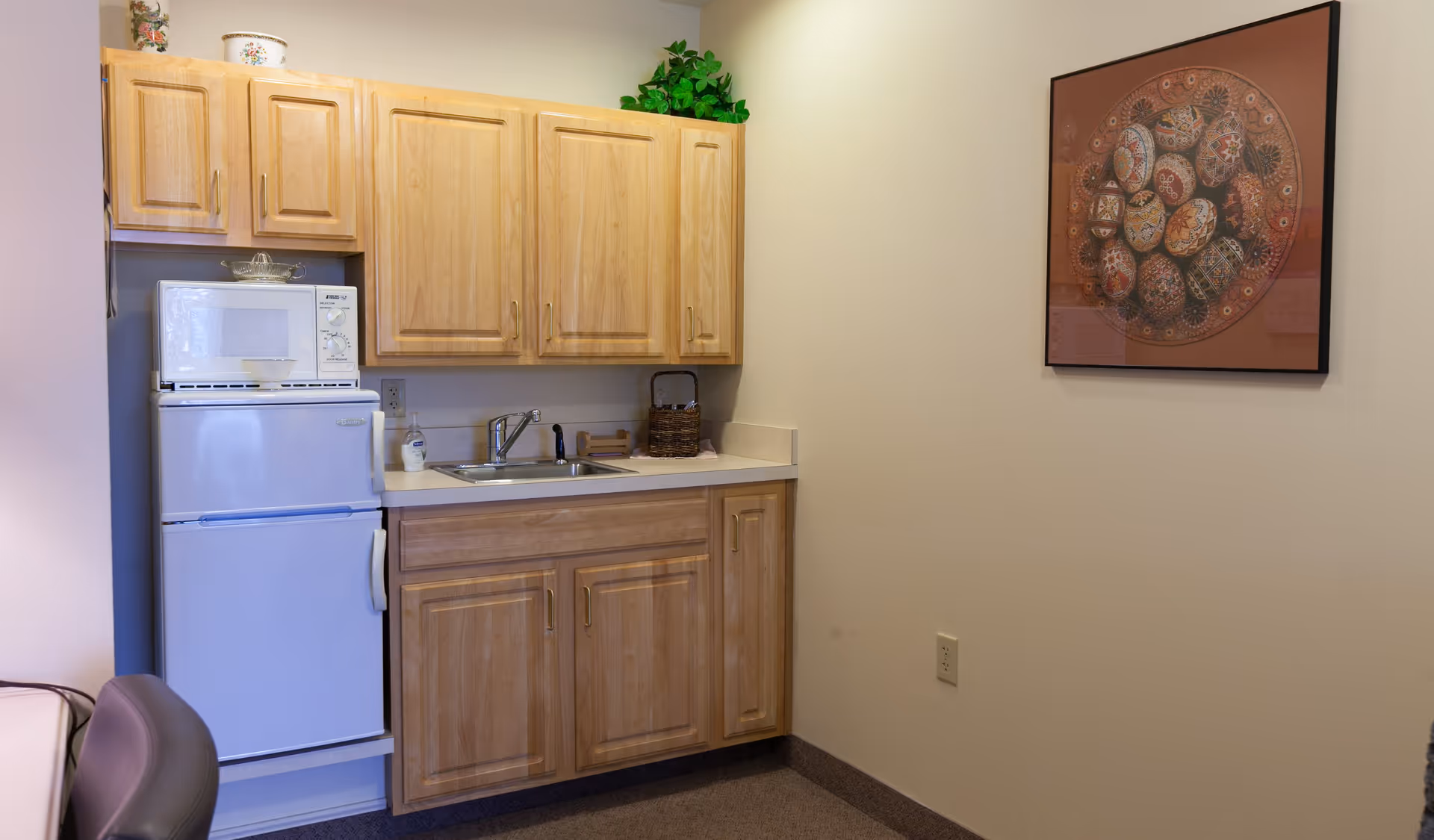 Small kitchenette area with light wood cabinets, a white mini refrigerator with a microwave on top, a sink with a faucet, a soap dispenser, and a basket on the countertop. A framed artwork featuring decorated eggs hangs on the adjacent wall.