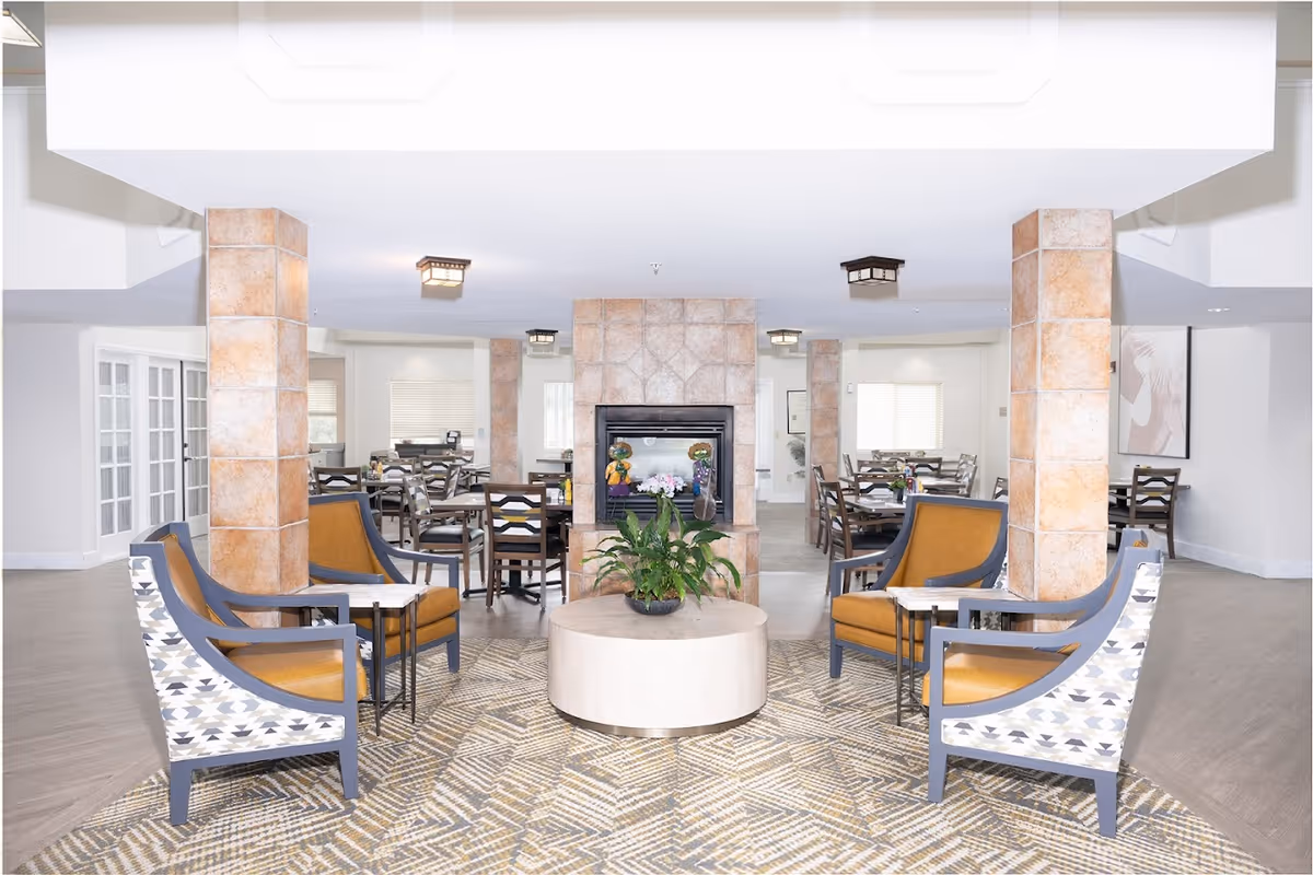 A bright and spacious senior living common area with a central fireplace surrounded by four tiled columns. The room features a patterned rug with four armchairs arranged around a round coffee table with a plant on it. In the background, there are multiple dining tables and chairs, and large windows letting in natural light.