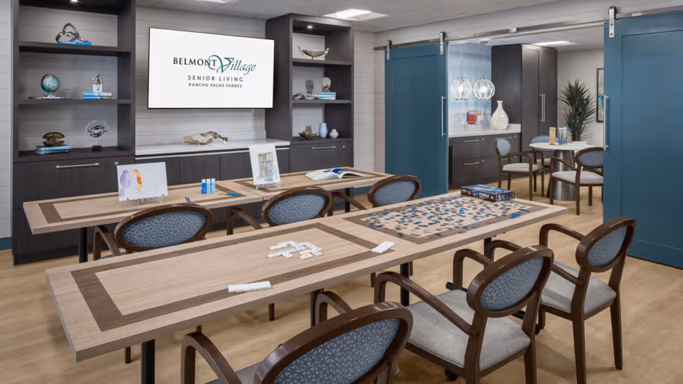 A well-lit activity room in a senior living facility with two long wooden tables and several chairs. One table has a partially completed jigsaw puzzle and dominoes, while the other has art supplies and paintings. In the background, there are dark wood shelves with decorative items and a large screen displaying the Belmont Village Senior Living Rancho Palos Verdes logo. To the right, there is a small round table with chairs and a cabinet with a beverage dispenser and decorative vases. The room features blue sliding doors and light wood flooring.