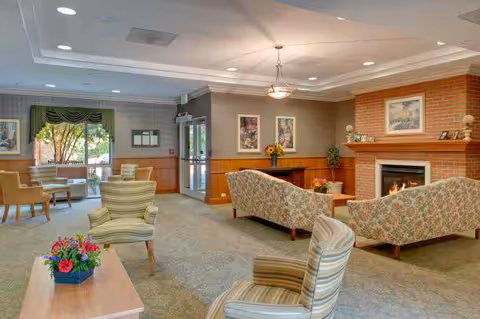 A cozy common room with upholstered chairs and sofas arranged around a fireplace and windows.