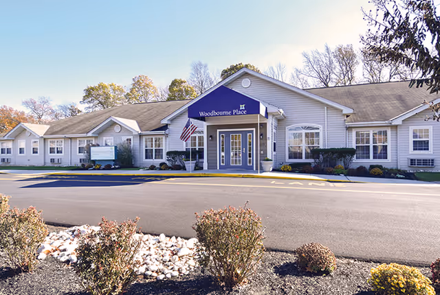 Front entrance of a single-story senior living facility with a blue awning reading 'Woodbourne Place', an American flag, and landscaped beds and a driveway in front.