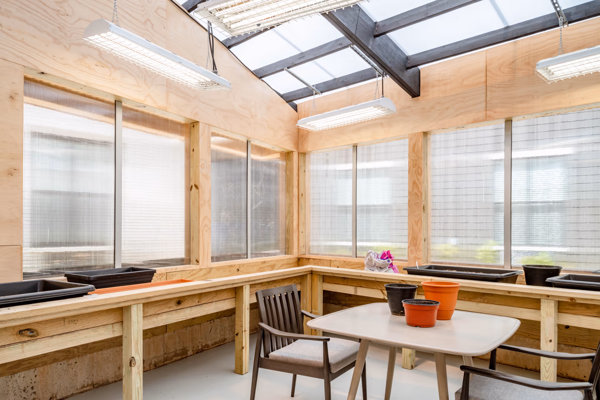 A bright indoor gardening room with wooden walls and large windows. There are several empty plant pots on a wooden counter and a table with chairs in the center. The ceiling has exposed beams and hanging fluorescent lights.