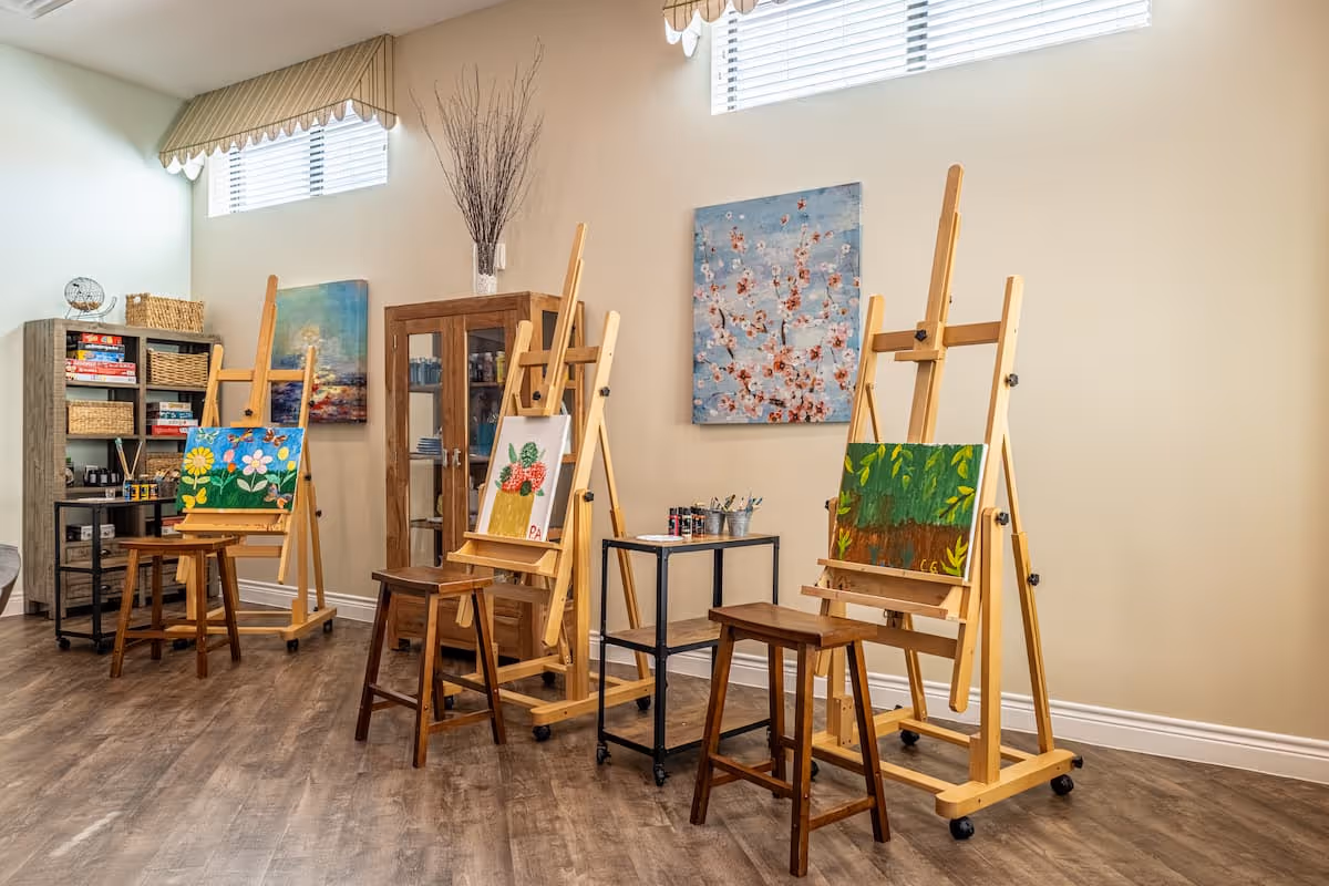 A bright art studio room with three wooden easels, each holding a colorful painting. There are three wooden stools in front of the easels. The room has light-colored walls, two high windows with blinds, and wooden flooring. A wooden cabinet and a bookshelf with board games and art supplies are visible in the background.