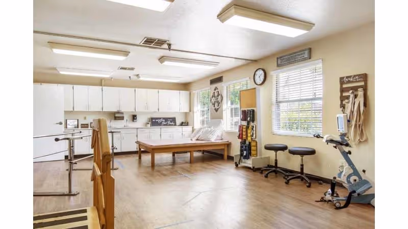 A spacious rehabilitation room with wooden flooring, parallel bars for walking exercises, a treatment table, exercise equipment, stools, and cabinets along the back wall. The room is well-lit with large windows and ceiling lights.
