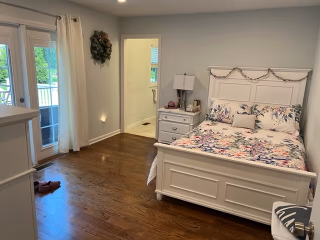 A cozy bedroom with a white wooden bed frame and floral bedding. There is a white nightstand with a lamp and some decorative items next to the bed. The room has wooden flooring and light-colored walls. A door leads to an adjoining bathroom, and there are glass doors with curtains that open to an outdoor area. A wreath hangs on the wall near the bathroom door.