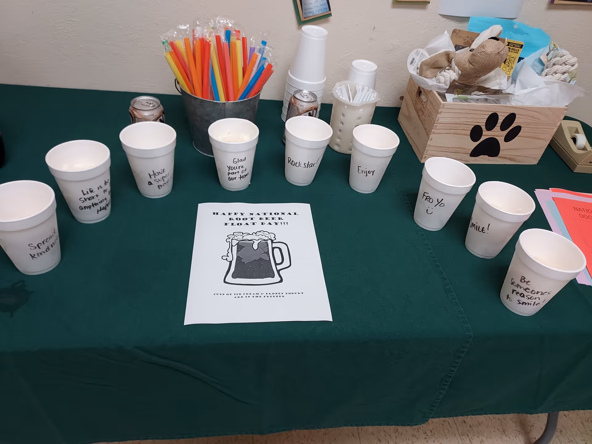 Table set up for National Root Beer Float Day with labeled disposable cups, colorful straws, and a flyer on a green tablecloth.