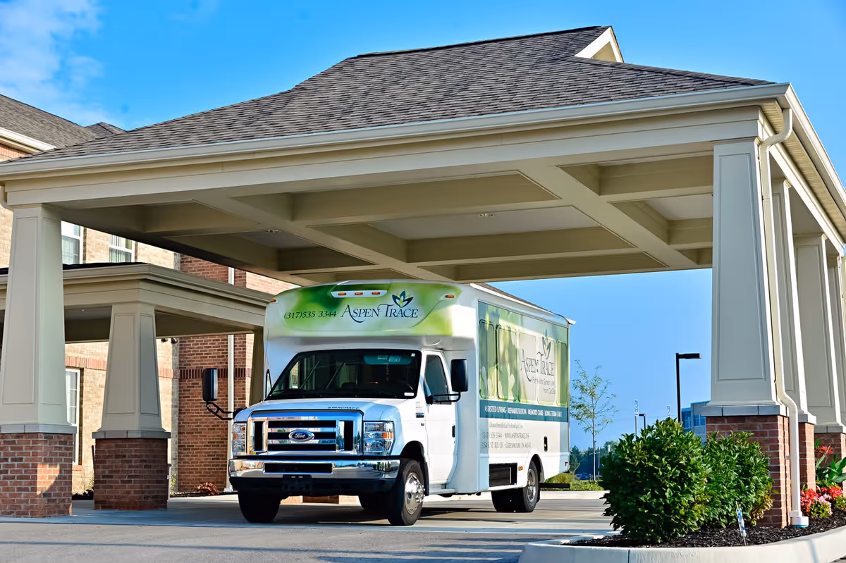 A white and green shuttle bus with Aspen Trace branding is parked under a covered entrance of a brick and beige building on a sunny day.