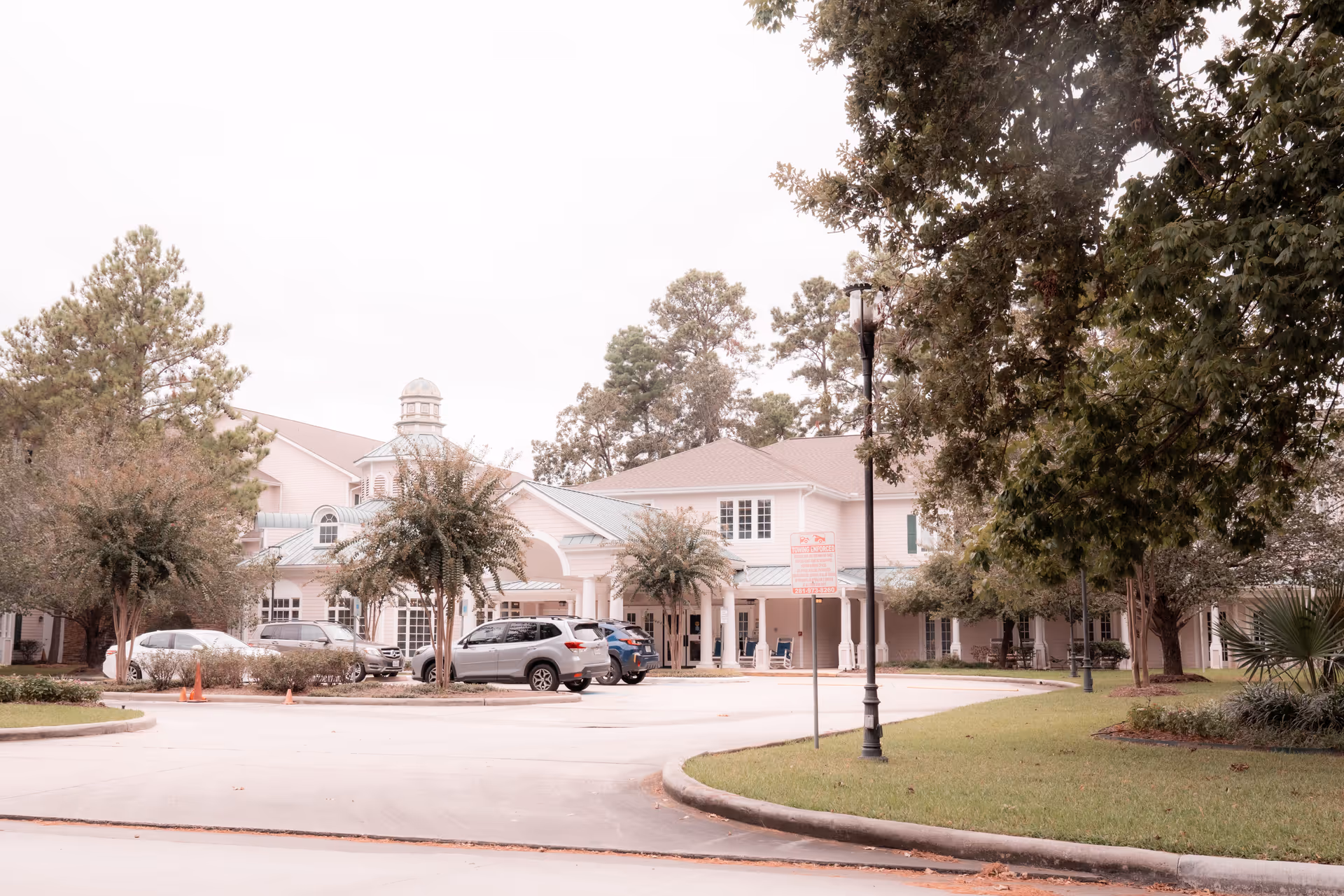 Exterior view of King's Preserve at Kingwood facility showing a large building with a light pink facade, green roof, and a cupola on top. Several cars are parked in front of the entrance, surrounded by trees and greenery under an overcast sky.